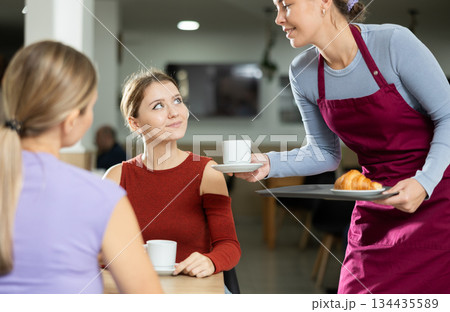 Girl waiter in apron presents coffee and croissants to restaurants guests. 134435589