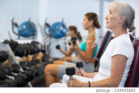 Slender senior woman sit and trains muscles of arms and back using dumb-bell shaped figure weighting Slender senior woman sit and trains muscles of arms and back using dumb-bell shaped figure weighting 134435883