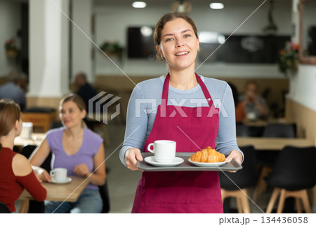 Female cafe employee carries coffee and croissants on tray Female cafe employee carries coffee and croissants on tray 134436058