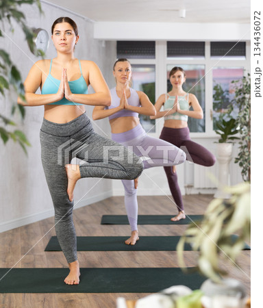 Diligent women practicing tree pose of yoga in light fitness room Diligent women practicing tree pose of yoga in light fitness room 134436072