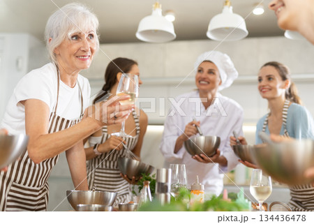 Aged woman holding wine and talking to other cooking course members whipping souce in bowls Aged woman holding wine and talking to other cooking course members whipping souce in bowls 134436478