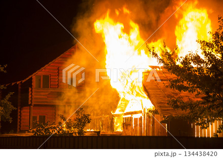Burning wooden house at night. Bright orange flames and dense smoke from under the tiled roof on dark sky, trees silhouettes and residential neighbor cottage background. Disaster and danger concept. 134438420