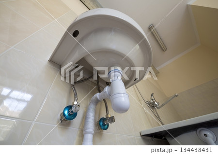 Bottom view of new white washbasin sink connected to sewer on background of bathroom light beige ceramic tiles. Professional plumbing and installation concept. Bottom view of new white washbasin sink connected to sewer on background of bathroom light beige ceramic tiles. Professional plumbing and installation concept. 134438431