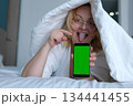 Middle aged woman under blanket sticking tongue out smiling holding smartphone with green screen showing potential advertising or digital content. 134441455