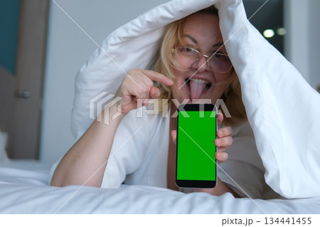 Middle aged woman under blanket sticking tongue out smiling holding smartphone with green screen showing potential advertising or digital content. Middle aged woman under blanket sticking tongue out smiling holding smartphone with green screen showing potential advertising or digital content. 134441455