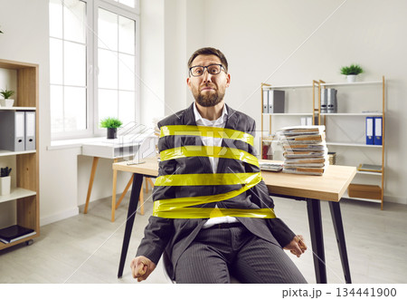 Sad man, trapped in office, wrapped with scotch tape, sitting by desk with paperwork 134441900