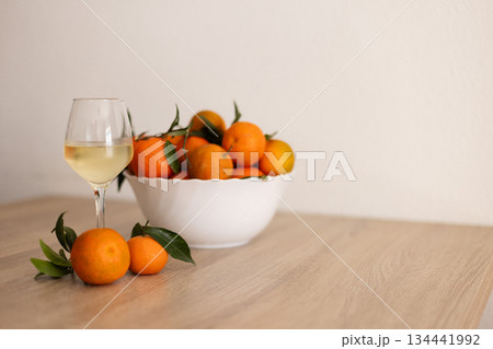 A glass of cold white wine next to tangerines on the wooden table. 134441992