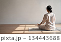 明るい日差しが差し込む清潔感のある室内でヨガマットの上に座り瞑想する女性の後ろ姿 134443688