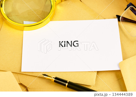 A magnifying glass, fountain pen, and glasses are placed around a card with the word King, laid out on yellow paper. Documents lie nearby, creating a scene of exploration and discovery 134444389