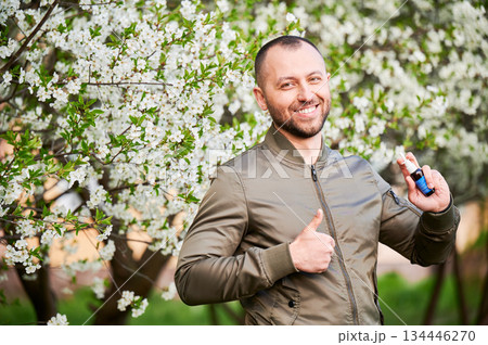 Man allergic using medical nasal drops, suffering from seasonal allergy at spring in blossoming garden. Handsome smiling man showing thumbs up near blooming tree outdoors. Spring allergy concept. 134446270