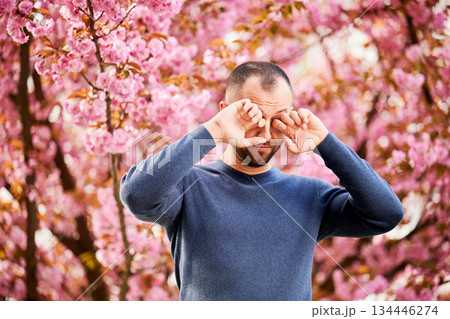 Man allergic suffering from seasonal allergy at spring in blossoming garden at springtime. Bearded man feeling itchy eyes in front of blooming tree. Spring allergy concept. 134446274