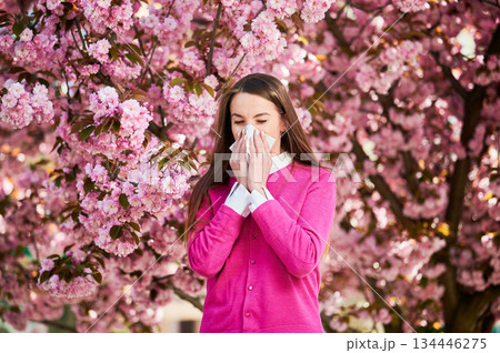 Woman allergic suffering from seasonal allergy at spring in blossoming garden at springtime. Woman sneezing and blowing nose using nasal handkerchief in front of blooming tree. Spring allergy concept. 134446275