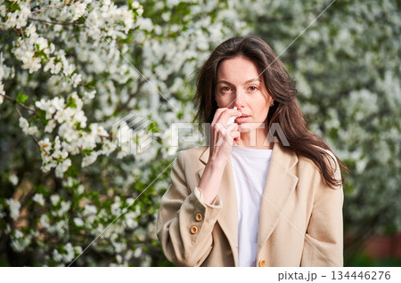 Woman allergic using medical nasal drops, suffering from seasonal allergy at spring in blossoming garden. Young woman treating runny nose in front of blooming tree outdoors. Spring allergy concept. 134446276
