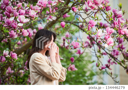 Woman allergic suffering from seasonal allergy at spring in blossoming garden at springtime. Young woman sneezing and blowing nose with nasal handkerchief in front of blooming tree. Allergy concept. 134446279