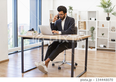 An Arabian businessman is working on his laptop while talking on the phone in his modern office An Arabian businessman is working on his laptop while talking on the phone in his modern office 134448070