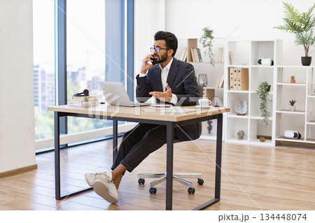 An Indian businessman is talking on the phone while working in his modern office setting An Indian businessman is talking on the phone while working in his modern office setting 134448074