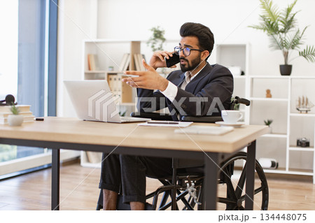 A businessman in a wheelchair is talking on the phone while working on his laptop in the office A businessman in a wheelchair is talking on the phone while working on his laptop in the office 134448075