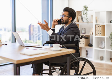 An Indian businessman in a wheelchair is talking on the phone in his office, looking serious An Indian businessman in a wheelchair is talking on the phone in his office, looking serious 134448076