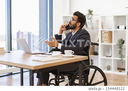 An Indian businessman in a wheelchair is talking on the phone while working in his office 134448122