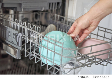 Hand placing colorful bowls in open dishwasher kitchen appliance interior Hand placing colorful bowls in open dishwasher kitchen appliance interior 134448487