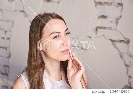 Caucasian young female applying skincare product on face with neutral expression 134448495