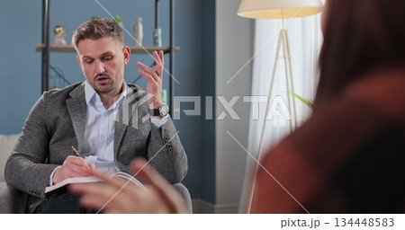 Man psychologist writing observation notes during therapy session with patient sitting beside in clinic room for mental health counseling support. 134448583