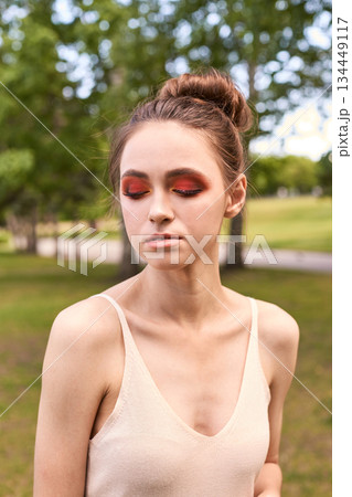 Young caucasian woman with red eye makeup outdoors in park setting with trees Young caucasian woman with red eye makeup outdoors in park setting with trees 134449117