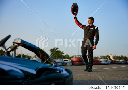 A kart racer stands proudly on the track, raising his helmet in a victorious gesture 134450445