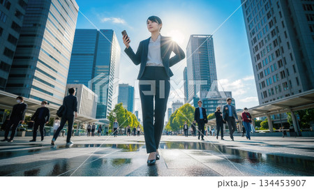 オフィス街をスマートフォン片手に歩くスーツ姿の若いビジネス女性　青空とビル群 134453907