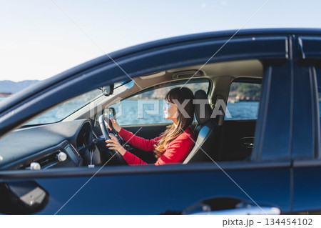 青空のもと運転席に乗る赤いニットのセーターを着た若いロングヘアの女性 134454102