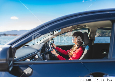青空のもと運転席に乗る赤いニットのセーターを着た若いロングヘアの女性 青空のもと運転席に乗る赤いニットのセーターを着た若いロングヘアの女性 134454103