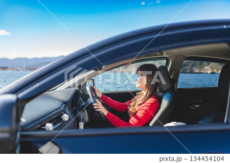 青空のもと運転席に乗る赤いニットのセーターを着た若いロングヘアの女性 134454104