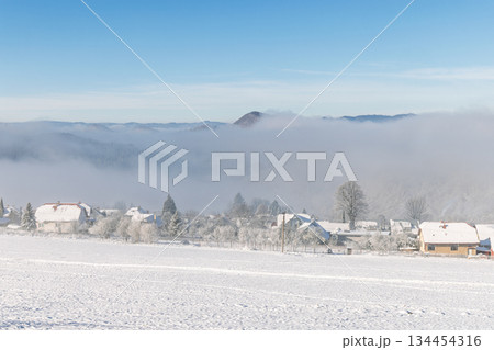Serene winter vista: A snow-covered village nestled beneath a blanket of fog, with hills visible in the distance under a clear blue sky. Peaceful winter scene. North of Slovakia, Europe. 134454316