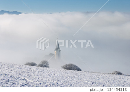 A serene winter landscape features a quaint church shrouded in fog, its steeple barely visible above the cloud line. The snow-covered field adds a touch of tranquility to the scene. A serene winter landscape features a quaint church shrouded in fog, its steeple barely visible above the cloud line. The snow-covered field adds a touch of tranquility to the scene. 134454318