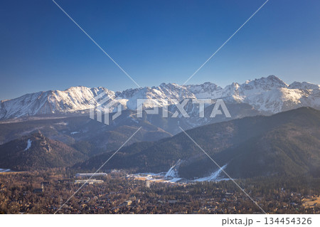 A scenic view of the mountains with snow-capped peaks, forests, and a town beneath a clear blue sky, creating a landscape. Zakopane town at the foot of the Tatra Mountains in Poland, Europe. 134454326