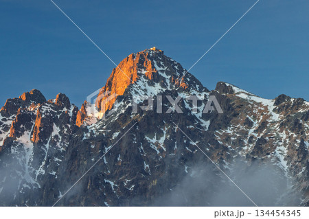 Lomnicky peak in High Tatras National Park, Slovakia, Europe. Golden hour bathes a majestic, snow-dusted mountain peak under clear blue skies, a serene and captivating alpine vista. 134454345