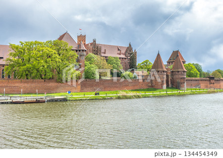 The Malbork Castle, a brick Gothic castle in northern Poland, Europe. Majestic medieval castle with red brick walls reflected in river under cloudy skies, showcasing historical architecture. 134454349