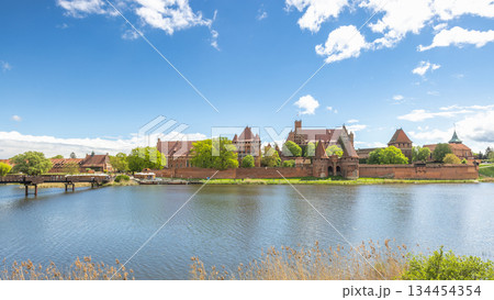 The Malbork Castle, a brick Gothic castle in northern Poland, Europe. Historical castle complex beside a reflective river under a bright sky with fluffy clouds on a sunny day. Medieval architecture. The Malbork Castle, a brick Gothic castle in northern Poland, Europe. Historical castle complex beside a reflective river under a bright sky with fluffy clouds on a sunny day. Medieval architecture. 134454354