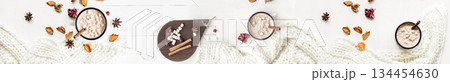 Panoramic top view of two mugs of hot chocolate with marshmallows, surrounded by cinnamon sticks, spices, and a warm white knitted scarf on a white wooden table. 134454630
