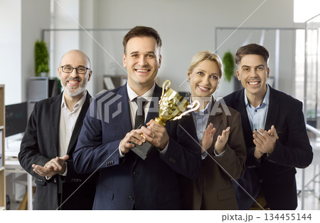 Happy business team applauding leader holding trophy in office after award win Happy business team applauding leader holding trophy in office after award win 134455144
