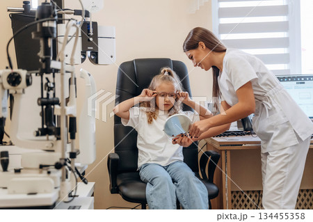 Holding mirror, looking at new eyewear. Little girl at the ophthalmologist clinic with doctor Holding mirror, looking at new eyewear. Little girl at the ophthalmologist clinic with doctor 134455358