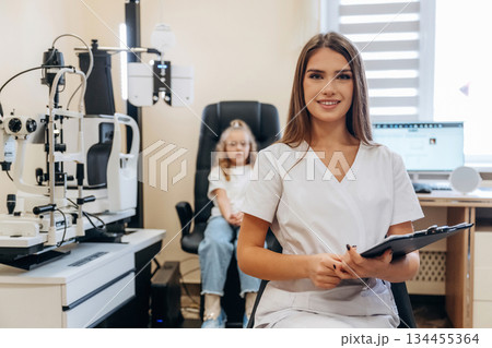 With notepad in hands. Little girl at the ophthalmologist clinic with doctor With notepad in hands. Little girl at the ophthalmologist clinic with doctor 134455364