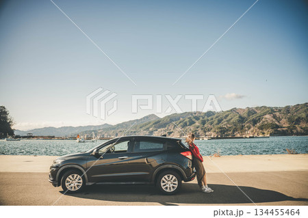 青空のもと冬の海で車に寄りかかって休憩するロングヘアの若い女性 134455464