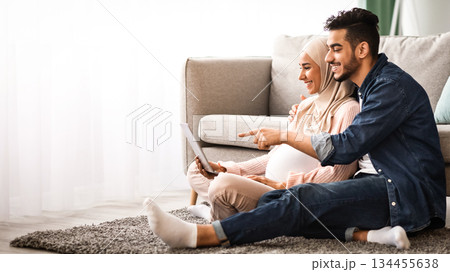 Happy islamic family man and pregnant woman using laptop together at home, cheerful middle eastern spouses making online shopping for future baby while relaxing with computer on floor in living room Happy islamic family man and pregnant woman using laptop together at home, cheerful middle eastern spouses making online shopping for future baby while relaxing with computer on floor in living room 134455638