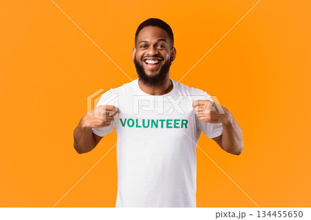 Volunteer's Portrait. Happy Black Activist Man Showing His T-Shirt With Volunteer Print Inscription On Chest Standing On Yellow Studio Background. Volunteering Job Concept 134455650