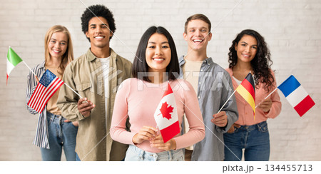 Young students of language school celebrate holiday. Happy millennial peoples of different nationalities have fun on study and hold little flags on brick white wall background, studio shot, free space Young students of language school celebrate holiday. Happy millennial peoples of different nationalities have fun on study and hold little flags on brick white wall background, studio shot, free space 134455713