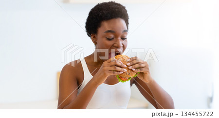 Cheat Meal Concept. Hungry Black Female Eating Big Burger Standing In Modern Kitchen At Home. Unhealthy Junk Food. Overeating And Weight Gain Problem, Eating Disorder And Nutrition Cheat Meal Concept. Hungry Black Female Eating Big Burger Standing In Modern Kitchen At Home. Unhealthy Junk Food. Overeating And Weight Gain Problem, Eating Disorder And Nutrition 134455722