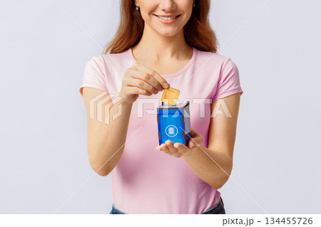 A millennial woman places a recyclable item into a small blue recycling bin at home, promoting eco-friendly practices. Her smile reflects a commitment to sustainability and environmental care. 134455726