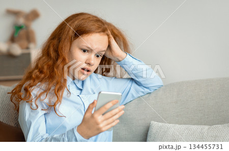 Shocked Surprised Young Teen Girl With Cellphone Reading Bad News Message Touching Head In Amazement Sitting On The Couch At Home. Confused Mobile Phone User, Call From Unknown Number 134455731