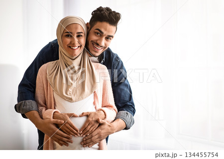 Portrait of happy pregnant religious muslim couple posing near window at home. Loving arab husband hugging his expecting wife in hijab from behind, making heart shaped sign on belly, copy space Portrait of happy pregnant religious muslim couple posing near window at home. Loving arab husband hugging his expecting wife in hijab from behind, making heart shaped sign on belly, copy space 134455754